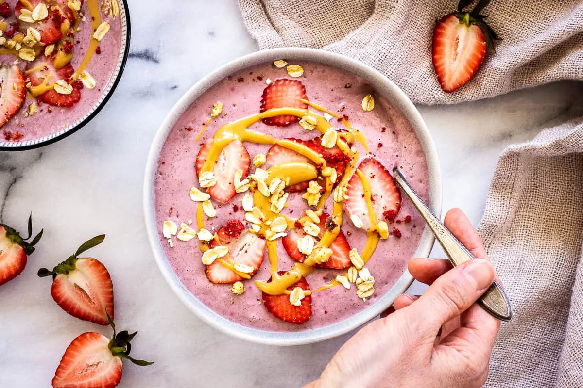 Overhead image shows a Strawberry Smoothie Bowl, decorated with strawberries, peanut butter and oats. A woman's hand dipping a silver spoon into the bowl is visible, as is a second bowl, a linen napkin and some strawberry halves.