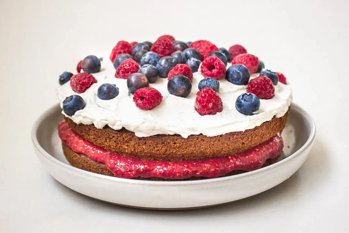 Vegan Gluten Free Cake. Cake is on a light grey plate on a white background and is decorated with raspberry jam, coconut whipped cream, blueberries and raspberries.