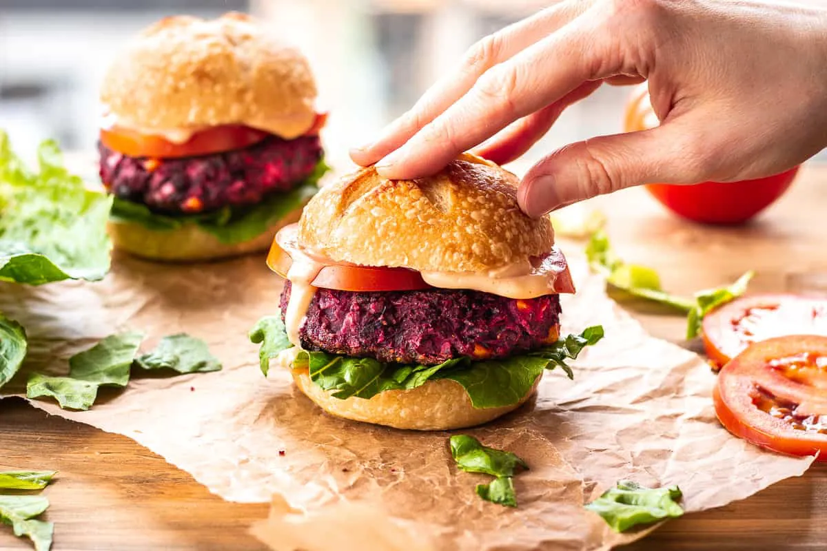Two Easy Beet & Black Bean Burgers on parchment paper. A woman's hand is lightly resting on top of the burger in the foreground. They are surrounded by lettuce leaves and tomato slices.