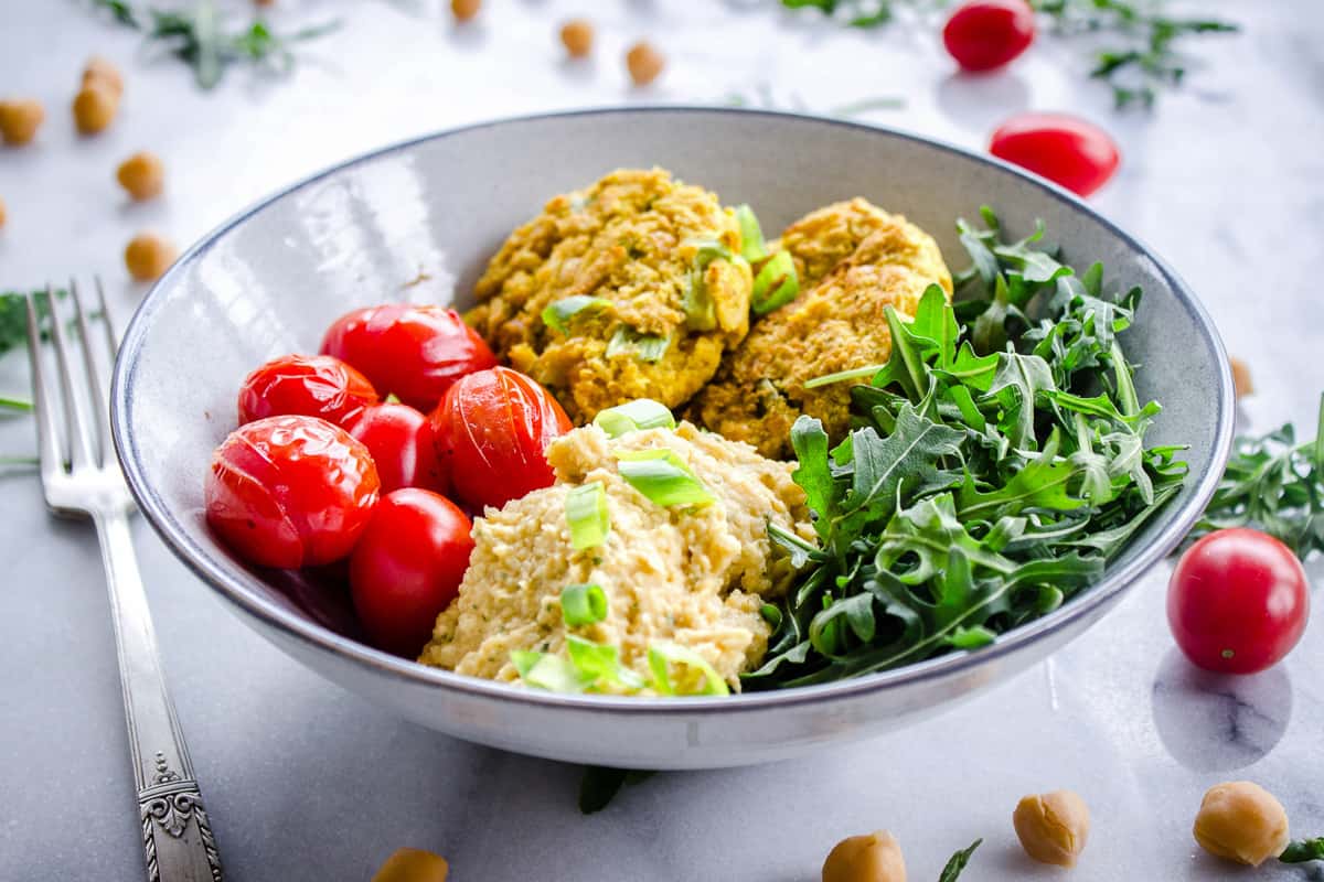Image shows Easy Vegan Chickpea Fritter Bowls on a white marble background. Bowl is filled with chickpea fritters, roasted cherry tomatoes, arugula and hummus. Tomatoes, chickpeas and arugula are scattered around the bowl.