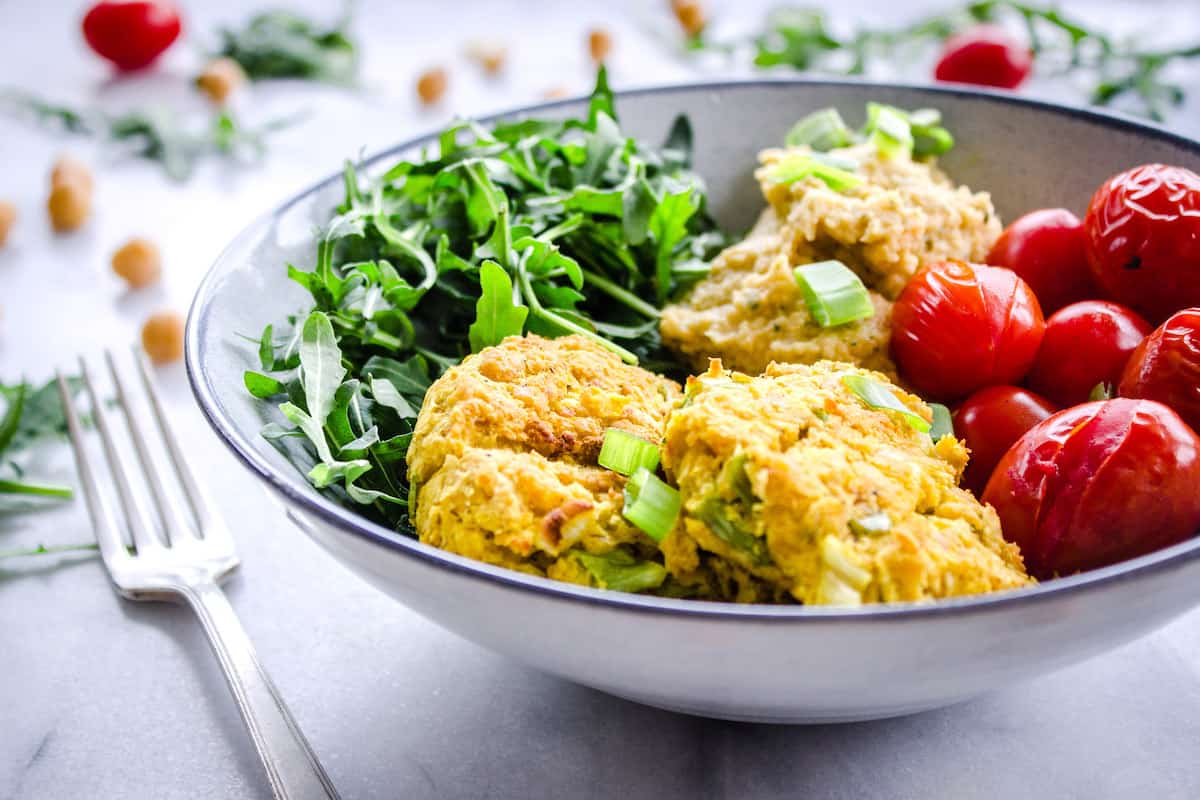 Close up image shows Easy Vegan Chickpea Fritter Bowls on a white marble background. Bowl is filled with chickpea fritters, roasted cherry tomatoes, arugula and hummus. Tomatoes, chickpeas and arugula are scattered around the bowl.