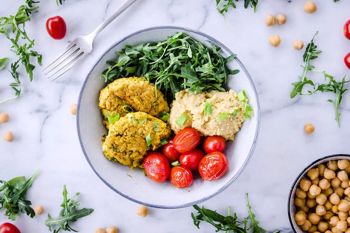 Overhead image shows Easy Vegan Chickpea Fritter Bowls on a white marble background. Bowl is filled with chickpea fritters, roasted cherry tomatoes, arugula and hummus. Tomatoes, chickpeas and arugula are scattered around the bowl.