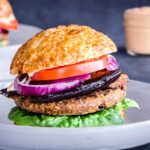 Image shows Smoky Eggplant Bean Burger on a grey plate layered with slices of tomato, red onion, smoky eggplant and lettuce. In the background is another burger and a jar of vegan garlic mayo.
