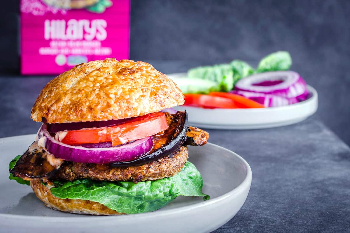Image shows Smoky Eggplant Bean Burger on a grey plate layered with slices of tomato, red onion, smoky eggplant and lettuce. In the background is box of Hilary's Adzuki Bean Burgers and a plate of sliced onions, tomatoes and lettuce.