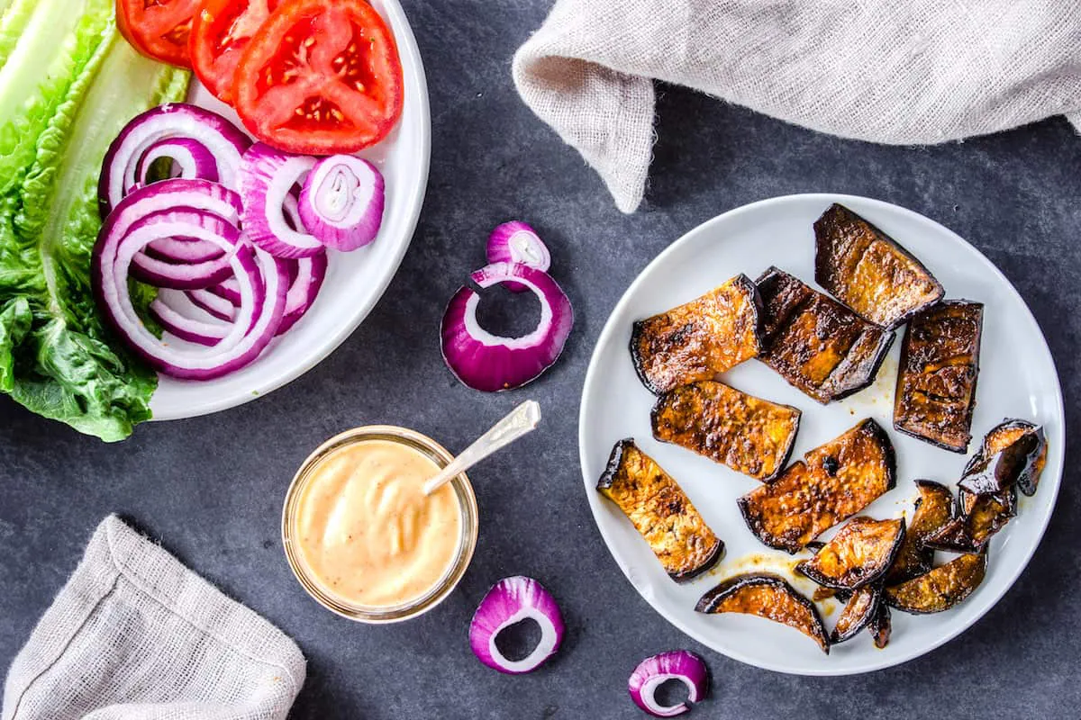 Overhead image shows ingredients for recipe on grey plates on a dark slate background. There is one plate of smoky eggplant slices, and one plate of sliced red onion, tomatoes and lettuce. There is also a jar of vegan garlic mayo and a linen napkin surrounding the plates.