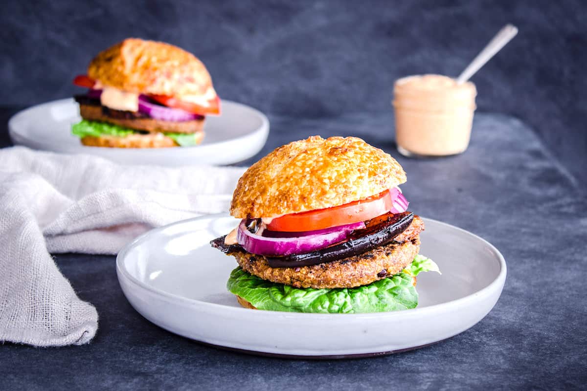 Image shows two Smoky Eggplant Bean Burgers on grey plates layered with slices of tomato, red onion, smoky eggplant and lettuce. A linen napkin is nearby. In the background is a jar of vegan garlic mayo.