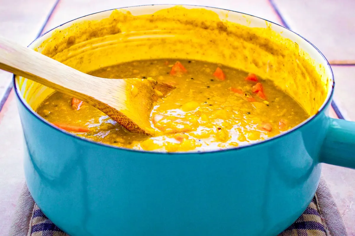 Image of Red Lentil Dhal in blue enamel saucepan on tiled surface. The dhal is a deep golden yellow with carrots in it. There is a wooden spoon in the saucepan, and it sits on a potholder.