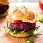 Close up image of Easy Beet & Black Bean Burger on parchment paper. The burger is surrounded by lettuce leaves and tomato slices, and there is a second burger in the background.