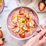 Strawberry Smoothie Bowl, decorated with strawberries, peanut butter and oats. A woman's hand dipping a silver spoon into the bowl is visible, as is a second bowl, a linen napkin and some strawberry halves.