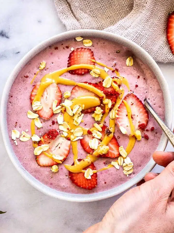 Strawberry Smoothie Bowl, decorated with strawberries, peanut butter and oats. A woman's hand dipping a silver spoon into the bowl is visible, as is a second bowl, a linen napkin and some strawberry halves.