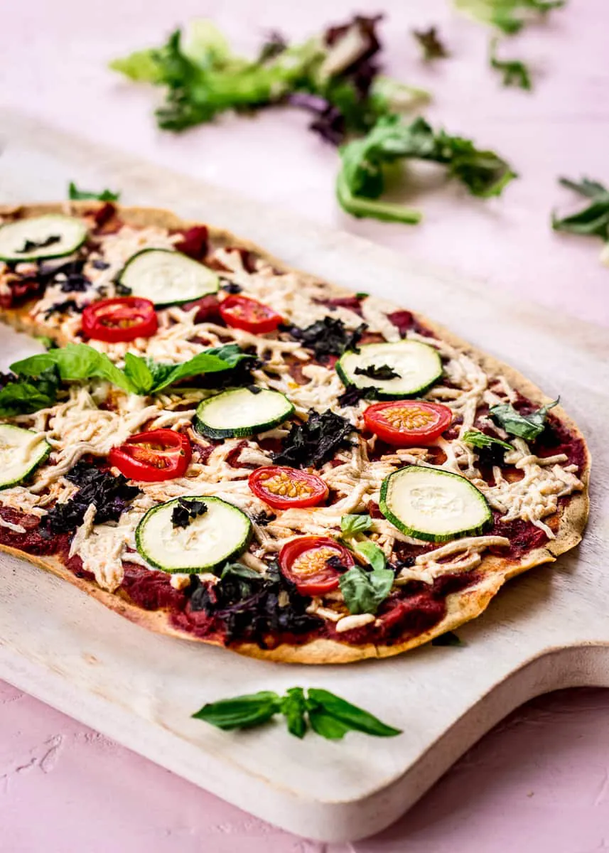 Close up image shows Chickpea Flour Pizza Crust on a white chopping board on a pink background. The pizza is topped with tomato sauce, vegan cheese, zucchini, tomatoes, spinach and basil, and there are salad leaves in the background.