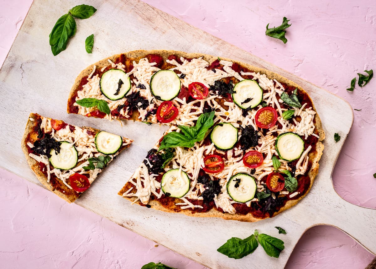 Overhead image shows Chickpea Flour Pizza Crust on a white chopping board on a pink background. The pizza is topped with tomato sauce, vegan cheese, zucchini, tomatoes, spinach and basil. A slice has already been cut from it.