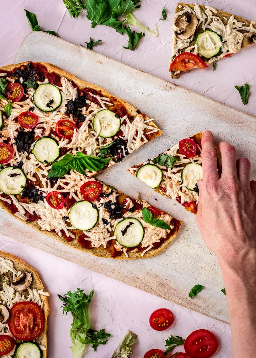 Overhead image shows Chickpea Flour Pizza Crust on a white chopping board on a pink background. The pizza is topped with tomato sauce, vegan cheese, zucchini, tomatoes, spinach and basil. A slice has already been cut from it, and a woman's hand is reaching for it. The pizza is surrounded by a pizza more slices of pizza, salad leaves and tomato slices.