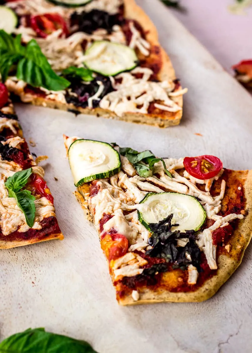 Close up image shows Chickpea Flour Pizza Crust on a white chopping board on a pink background. The pizza is topped with tomato sauce, vegan cheese, zucchini, tomatoes, spinach and basil. A slice has already been cut from it.