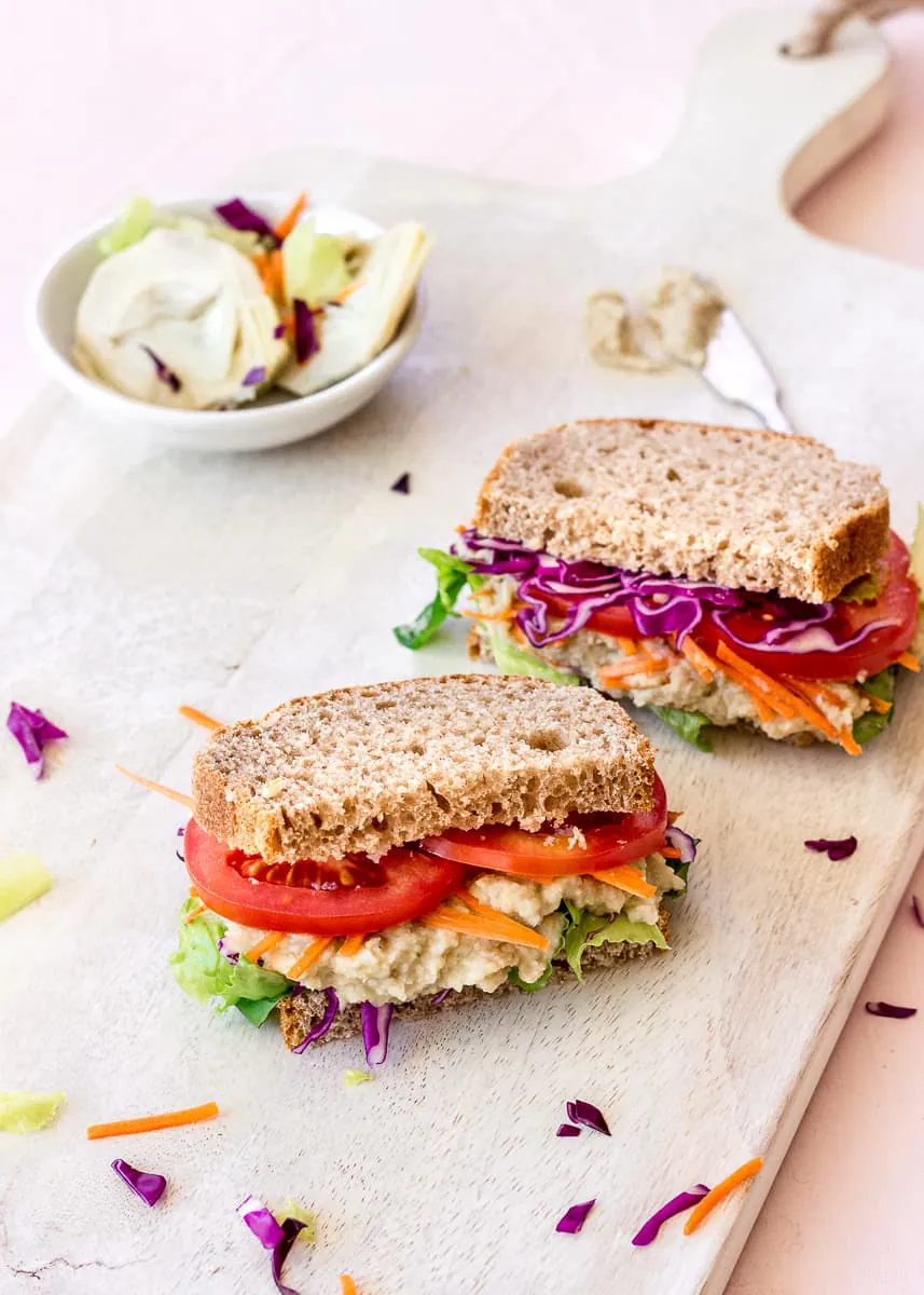 Side image of two sandwiches on a white wooden chopping board. The sandwiches are filled with brightly coloured vegetables and spread, and behind them is a dish of artichokes and other vegetables.