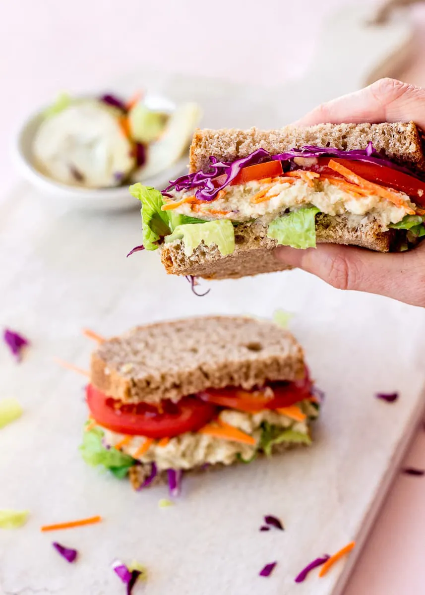 Image of a woman's hand holding a White Bean & Artichoke Vegan Sandwich. In the background there is a dish of artichokes and another sandwich.