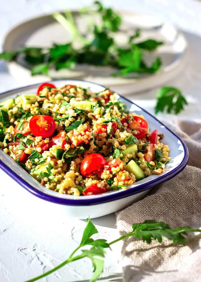 Image shows one bowl of Gluten-free Millet Tabbouleh on a white plaster background. The tabbouleh contains tomatoes, parsley and cucumber. Behind it is a plate with scattered parsley leaves.