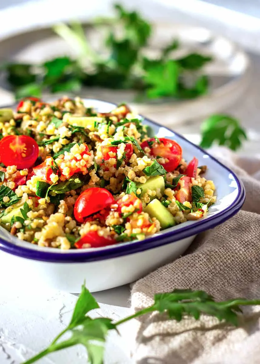 Image shows one bowl of Gluten-free Millet Tabbouleh on a white plaster background. The tabbouleh contains cucumber, parsley and cucumber. Behind it is a plate with scattered parsley leaves.
