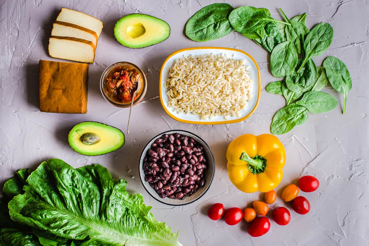 Overhead shot of ingredients on a grey plaster background, including avocado, brown rice, smoked tofu, bell pepper, black beans and spinach.