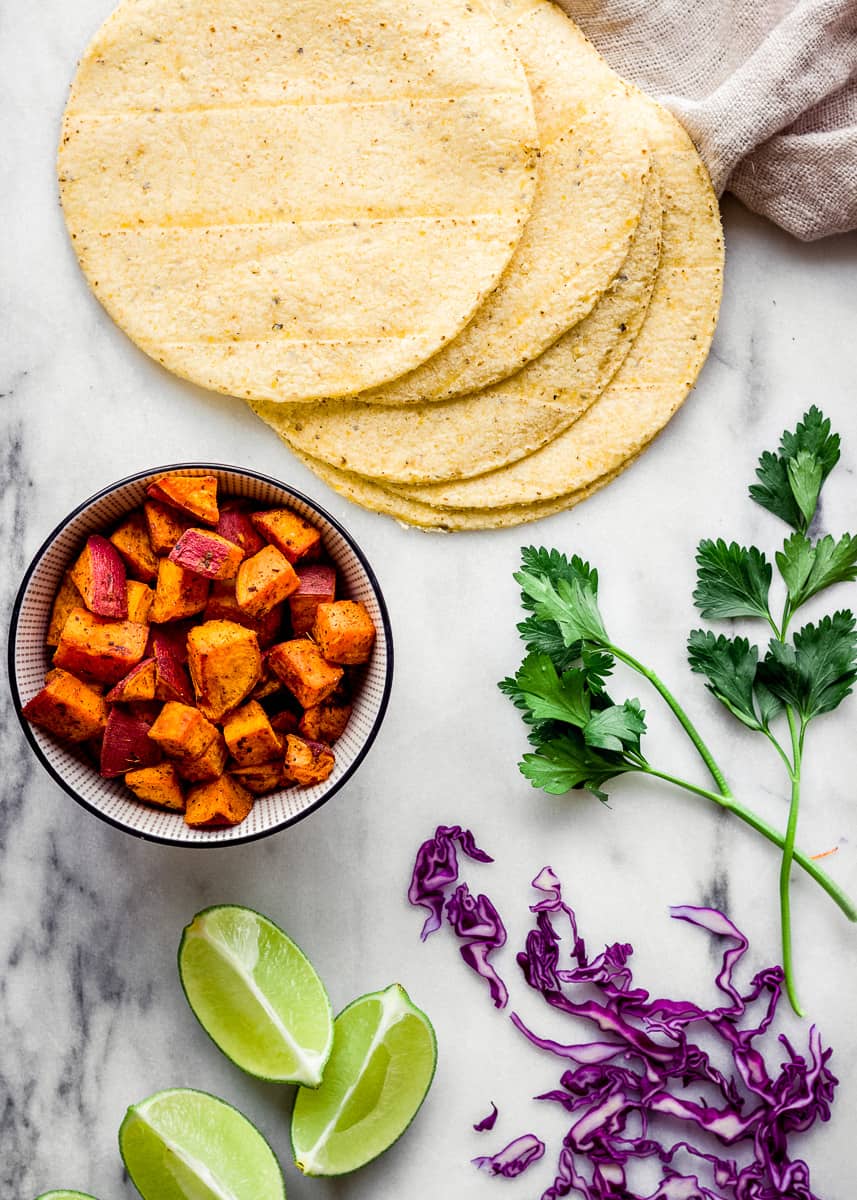 Overhead image of ingredients on a marble surface, including a bowl of roasted yams, red cabbage, corn tortillas, cilantro and lime wedges.