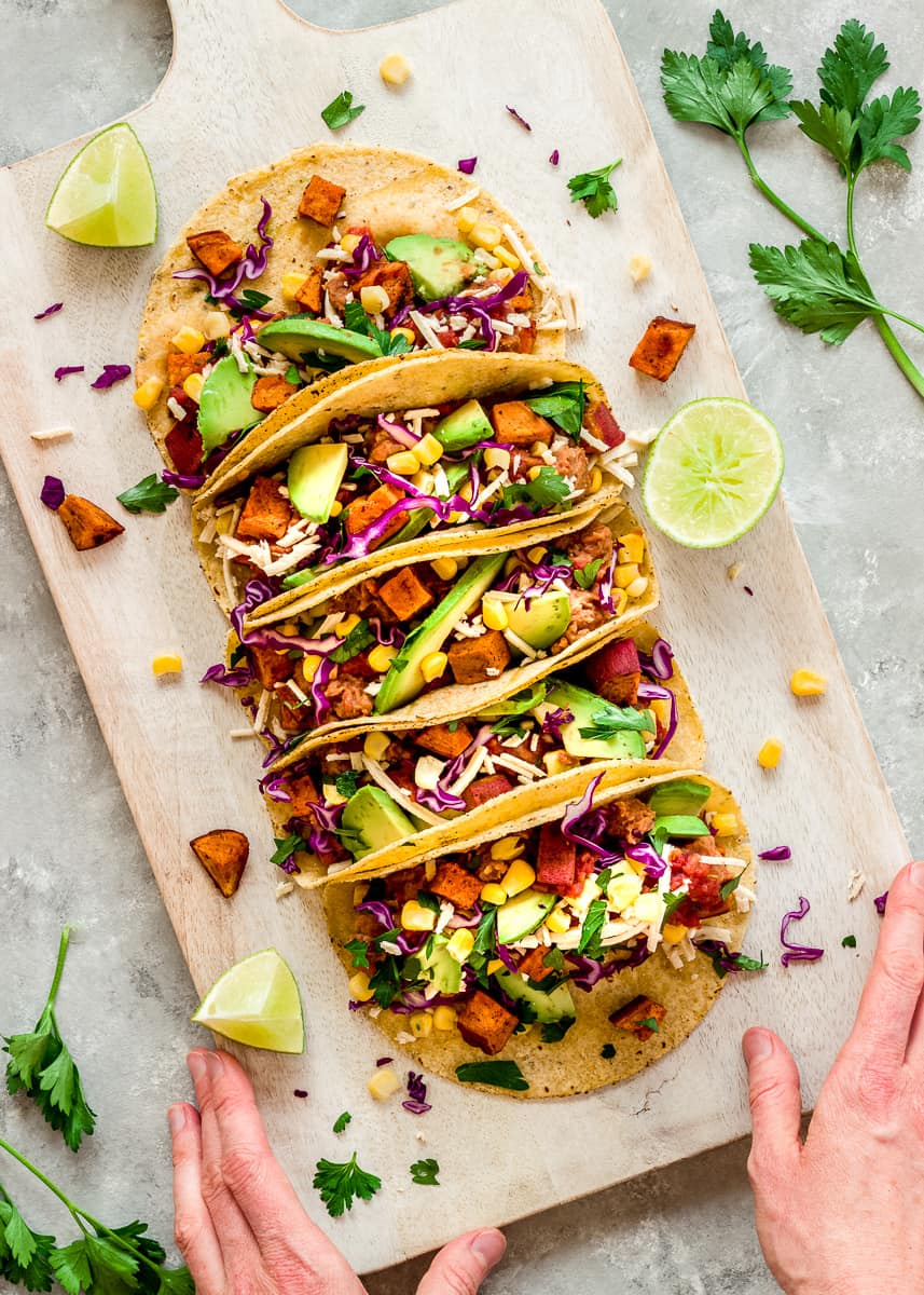 Overhead image of Roasted Sweet Potato Tacos on a white wooden chopping board. The tacos are decorated with red cabbage, avocado and sweetcorn. A woman's hands hold the chopping board and there are wedges of lime and cilantro leaves surrounding the tacos.