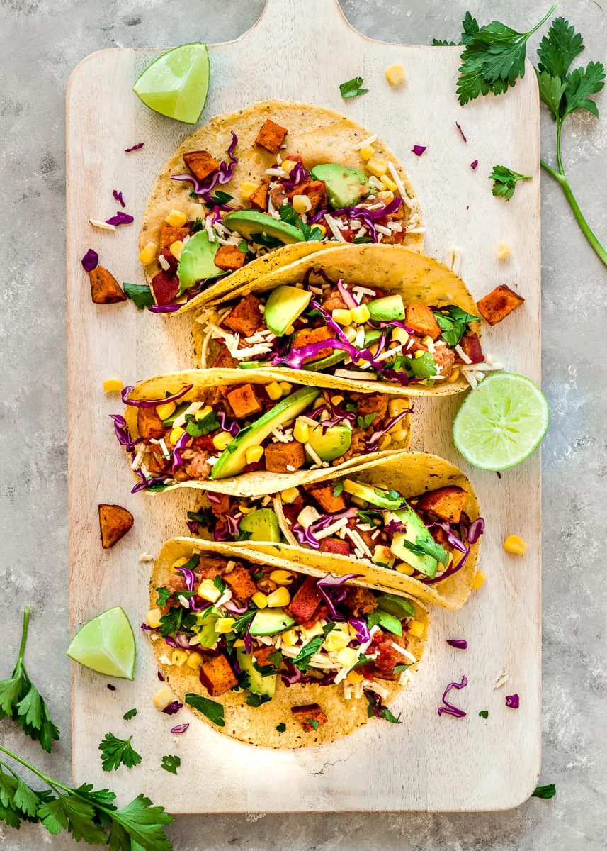 Overhead image of Roasted Sweet Potato Tacos on a white wooden chopping board. The tacos are decorated with red cabbage, avocado and sweetcorn. There are wedges of lime and cilantro leaves surrounding the tacos.