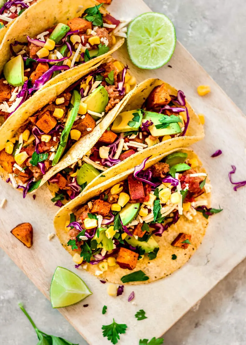 Partial overhead image of Roasted Sweet Potato Tacos on a white wooden chopping board. The tacos are decorated with red cabbage, avocado and sweetcorn. There are wedges of lime and cilantro leaves surrounding the tacos.
