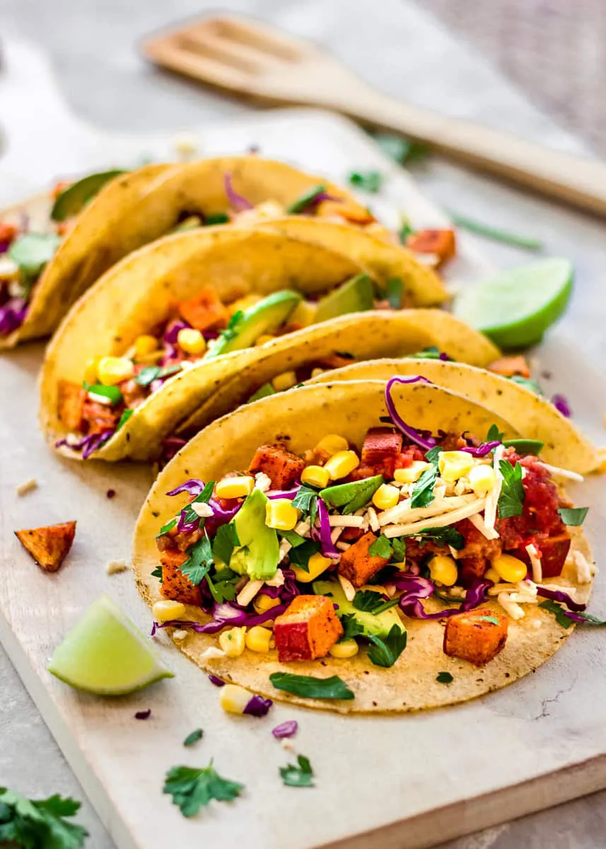 Side image of Roasted Sweet Potato Tacos on a white wooden chopping board at an angle. The tacos are decorated with red cabbage, avocado and sweetcorn. There is a wooden spatula in the background and there are wedges of lime and cilantro leaves surrounding the tacos.