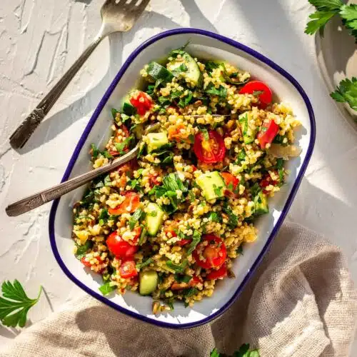 Overhead shot of one white enamel bowl of Gluten-free Millet Tabbouleh on a white plaster background surrounded by forks, parsley leaves and a napkin. The tabbouleh contains tomatoes, parsley and cucumber.