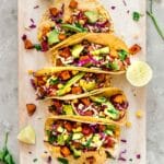 Overhead image of Roasted Sweet Potato Tacos on a white wooden chopping board. The tacos are decorated with red cabbage, avocado and sweetcorn. A woman's hands hold the chopping board and there are wedges of lime and cilantro leaves surrounding the tacos.