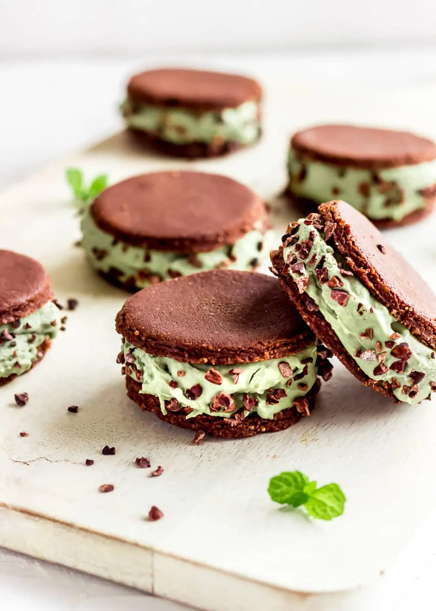 Vegan ice cream sandwiches on a white chopping board decorated with mint leaves. The sandwiches contain layers of mint ice cream between raw chocolate cookies and are decorated with cacao nibs.