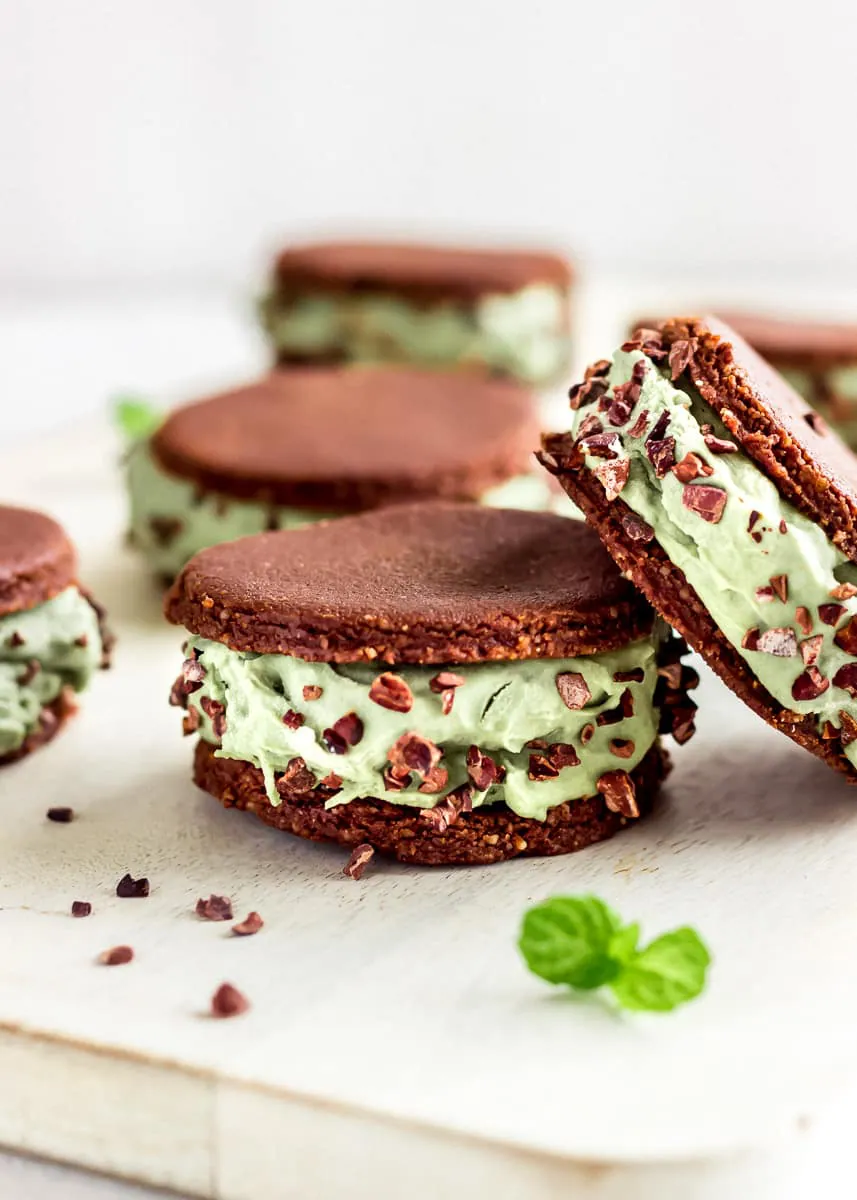 Close up view of vegan ice cream sandwiches against a white background decorated with mint leaves. The sandwiches contain layers of mint ice cream between raw chocolate cookies and are decorated with cacao nibs.