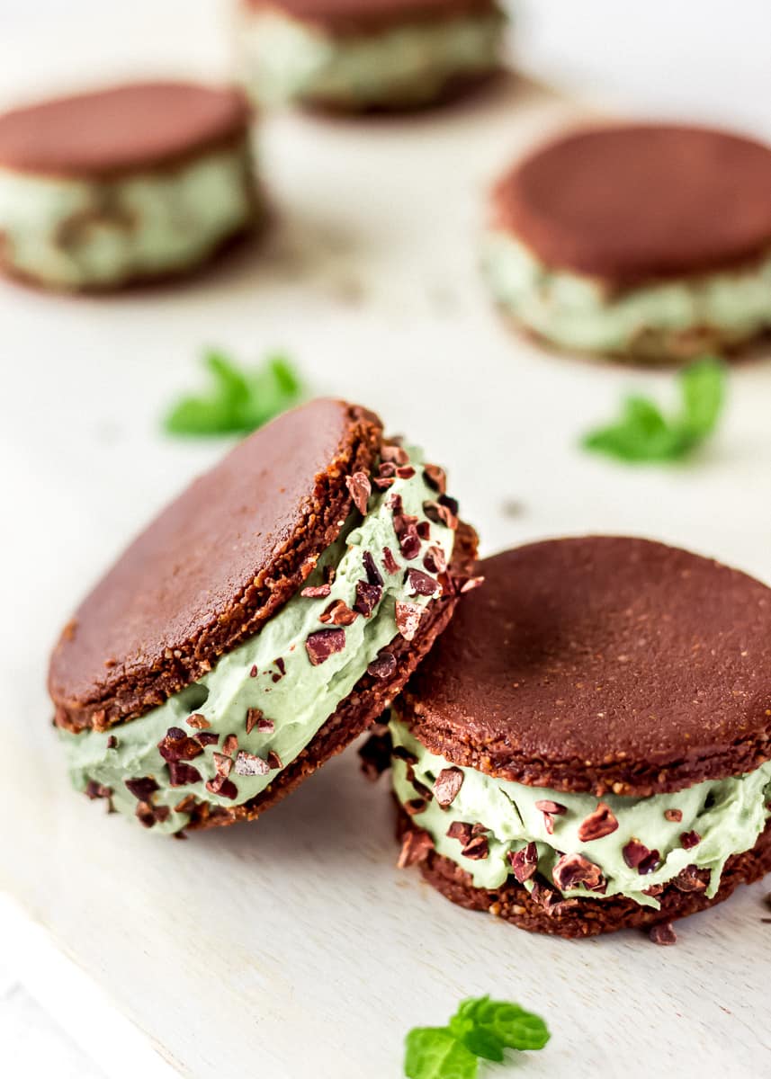 Close up view of vegan ice cream sandwiches against a white background decorated with mint leaves. The sandwiches contain layers of mint ice cream between raw chocolate cookies and are decorated with cacao nibs.