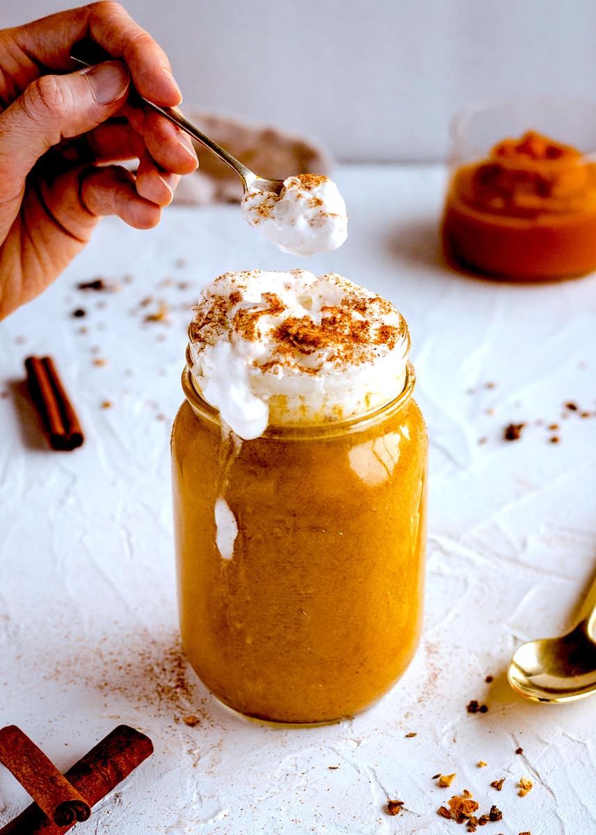 Tall vegan drink in a mason jar against a white textured background. It is in a glass mason jar and is topped with coconut whipped cream and pumpkin spice. A spoon with whipped cream on is being held by a woman's hand above the drink. Surrounding it are cinnamon sticks, a golden spoon, a linen napkin and a jar of pumpkin puree. 