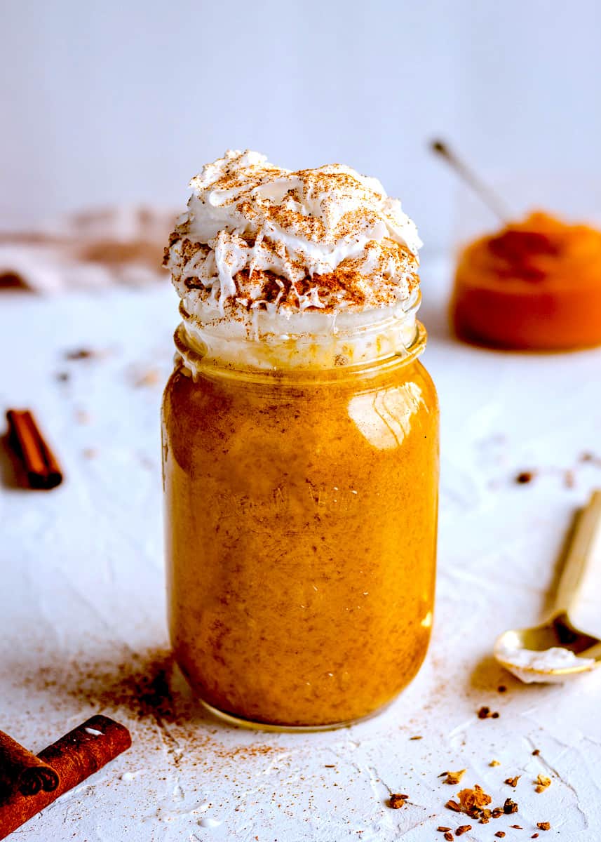 Tall vegan drink in a mason jar against a white textured background. It is in a glass mason jar and is topped with coconut whipped cream and pumpkin spice. Surrounding it are cinnamon sticks, a golden spoon, a linen napkin and a jar of pumpkin puree. 