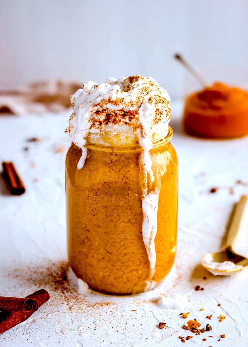 5-Ingredient Pumpkin Spice Latte against a white textured background. It is in a glass mason jar and is topped with coconut whipped cream and pumpkin spice. Surrounding it are cinnamon sticks, a golden spoon and a jar of pumpkin puree.