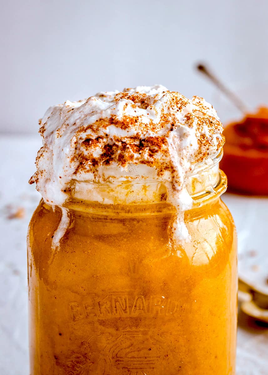 Close up of vegan drink topped with whipped cream and pumpkin spice. The drink is a deep golden colour and a jar of pumpkin is just visible in the background.