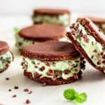 Vegan ice cream sandwiches against a white background decorated with mint leaves. The sandwiches contain layers of mint ice cream between raw chocolate cookies and are decorated with cacao nibs.