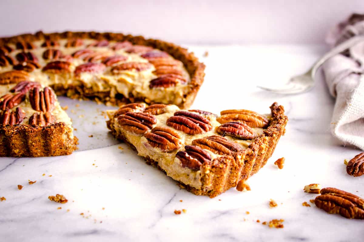 Image shows a slice of Easy Vegan Pecan Pie sitting on a white marble background next to the rest of the pie. It is decorated with concentric circles of pecans and surrounding it are a beige linen napkin, a silver fork and pecan halves.