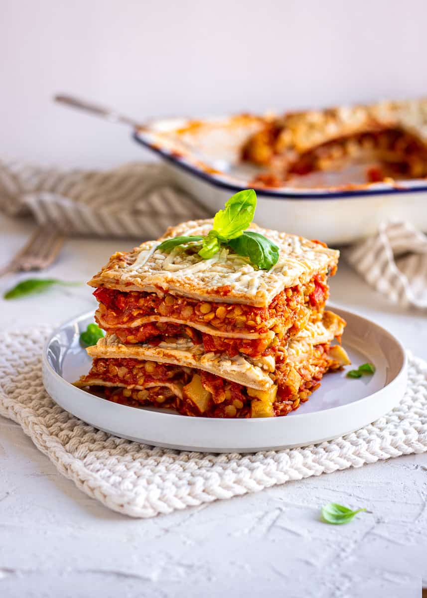 Image shows a large wedge of red lentil lasagna on a white plate. Behind it is a large dish of lasagna.