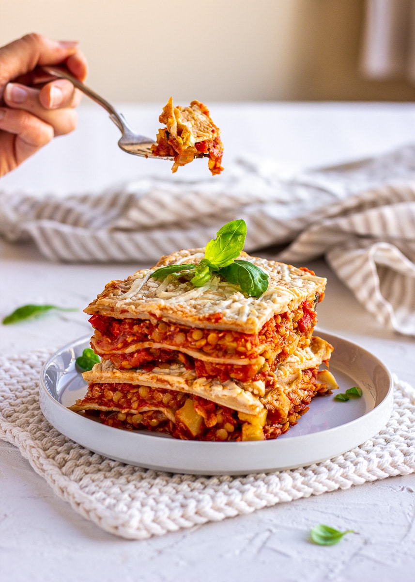 Image shows a large wedge of red lentil lasagna on a white plate. A woman's hand can be seen lifting a forkful of the lasagna.