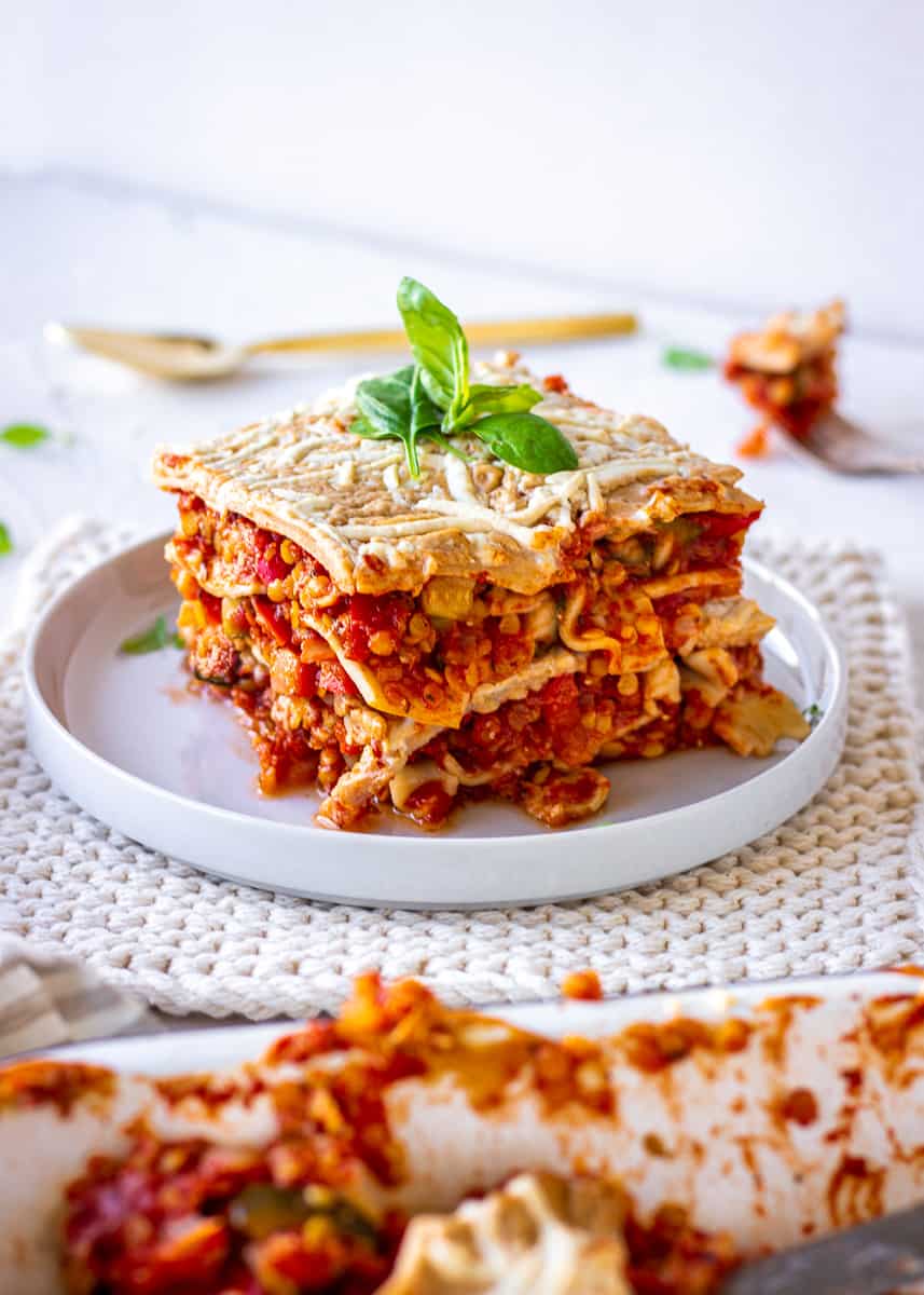 A thick slice of Easy Weeknight Vegan Lasagna is displayed on a white plate. It is topped with basil leaves and the plate is sitting on a white potholder. In the foreground of the image is the oven dish of lasagne.