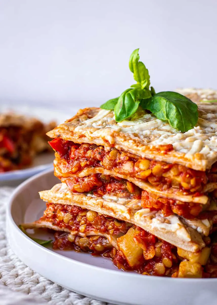 Close up of vegan lasagna, showing layers of red lentils and tomato sauce, vegetables, pasta and vegan cheese.