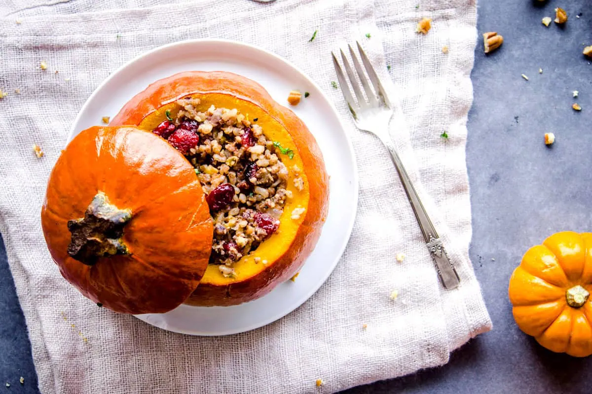 Overhead shot of Stuffed Pumpkin, showing a pumpkin with its lid off, stuffed with cranberries, rice and nuts on a white plate. The plate is on a folded linen napkin which is sitting on a grey table. Around it is a smaller pumpkin, silver cutlery and a dish of pecans.