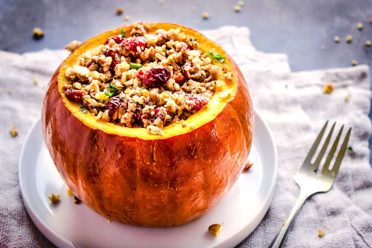 Closeup shot of Stuffed Pumpkin, showing a pumpkin without its lid, stuffed with cranberries, rice and nuts on a white plate. The plate is on a folded linen napkin which is sitting on a grey table. Surrounding it is silver cutlery and pecan crumbs.