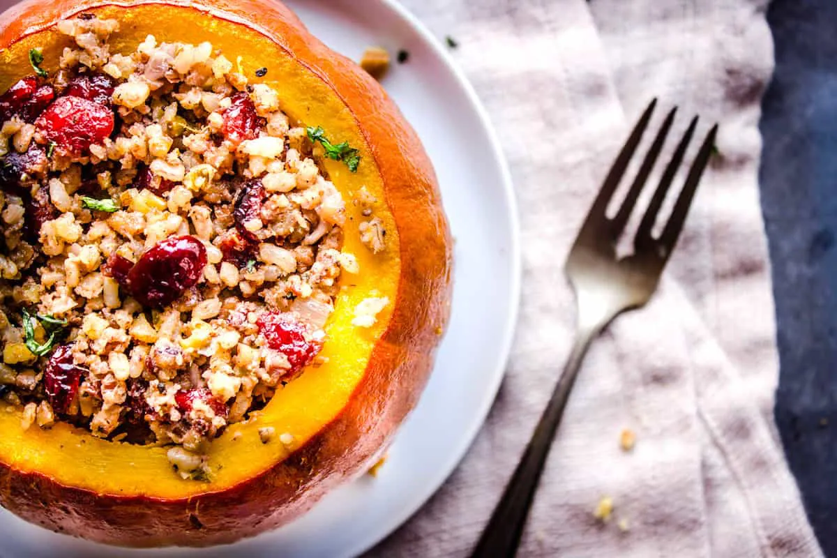 Closeup overhead shot of Stuffed Pumpkin, showing a pumpkin without its lid, stuffed with cranberries, rice and nuts on a white plate. The plate is on a folded linen napkin which is sitting on a grey table. Surrounding it is a silver fork and pecan crumbs.