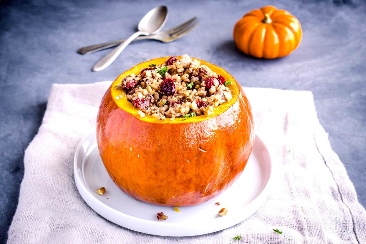 Side shot of a pumpkin without its lid sitting on a beige linen napkin. The pumpkin is stuffed with cranberries, rice and nuts. Surrounding it is silver cutlery and a smaller pumpkin.