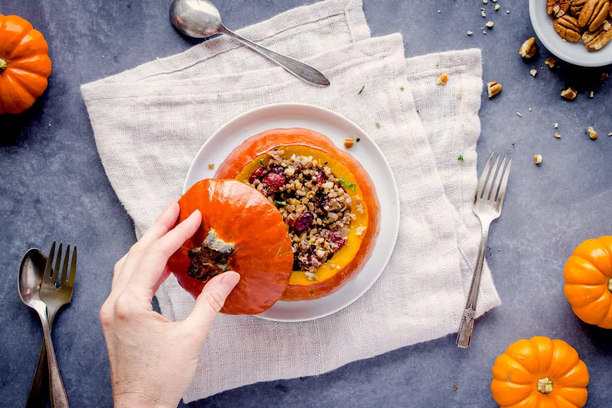 Overhead shot of Stuffed Pumpkin, showing a female hand holding the lid of a pumpkin stuffed with cranberries, rice and nuts on a white plate. The plate is on a folded linen napkin which is sitting on a grey table. Around it are smaller pumpkins, silver cutlery and a dish of pecans.
