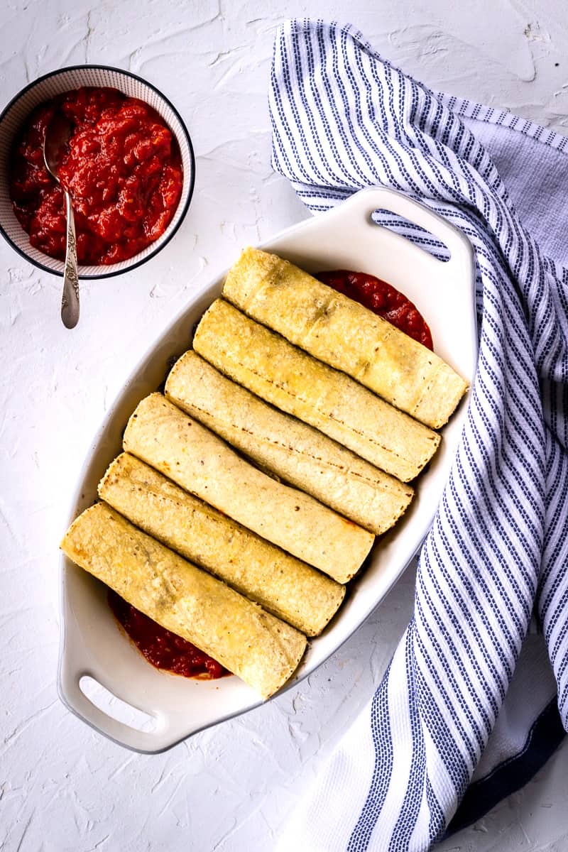 Overhead image of corn tortillas rolled neatly on top of a tomato sauce in a white casserole dish. A dish of enchilada sauce sits next to it, and the is a blue and white striped tea towel to one side.