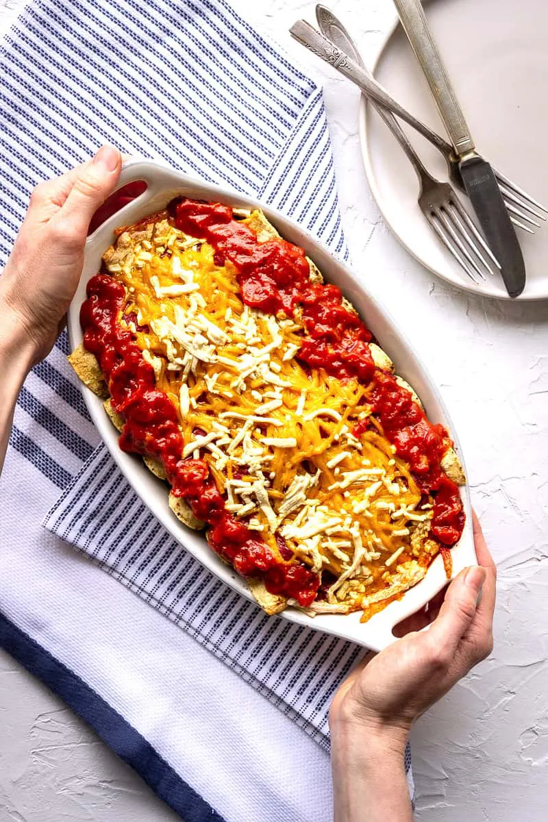 A woman's hands hold a dish of vegan enchiladas decorated with vegan cheese.